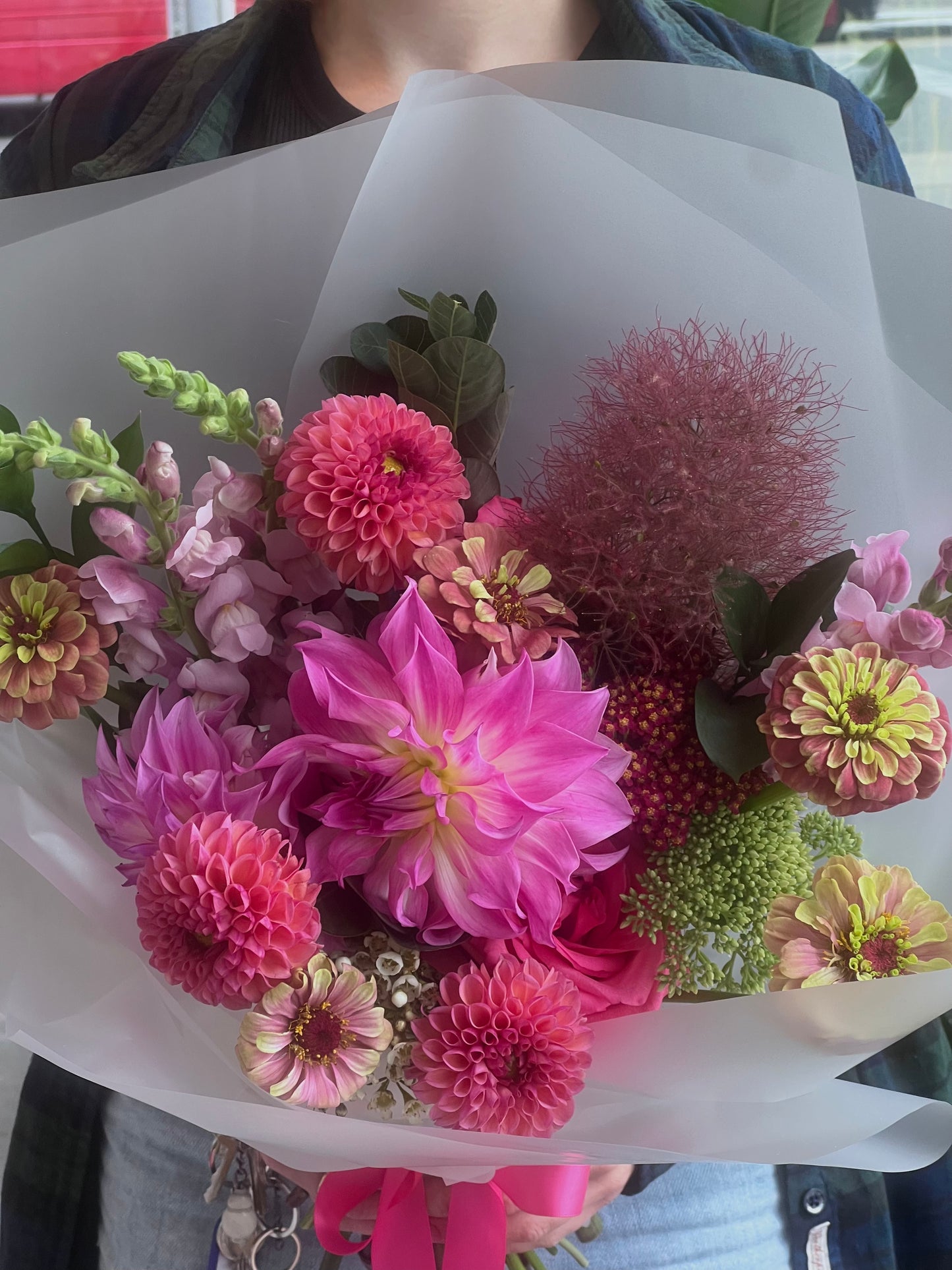 A vibrant mixed flower bouquet wrapped in white paper, held by a person wearing a blue denim jacket. The bouquet features bright pink dahlias, magenta and peach zinnias, light pink stock, smokebush and green foliage.