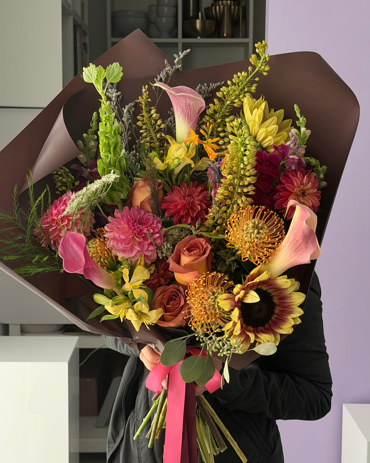 A large, fall flower bouquet wrapped in dark brown paper and tied with a coral ribbon, Held by a person wearing a black jacket. This bouquet features a variety of golden hour fall flowers including pink, yellow, orange, and red blooms with green foliage.