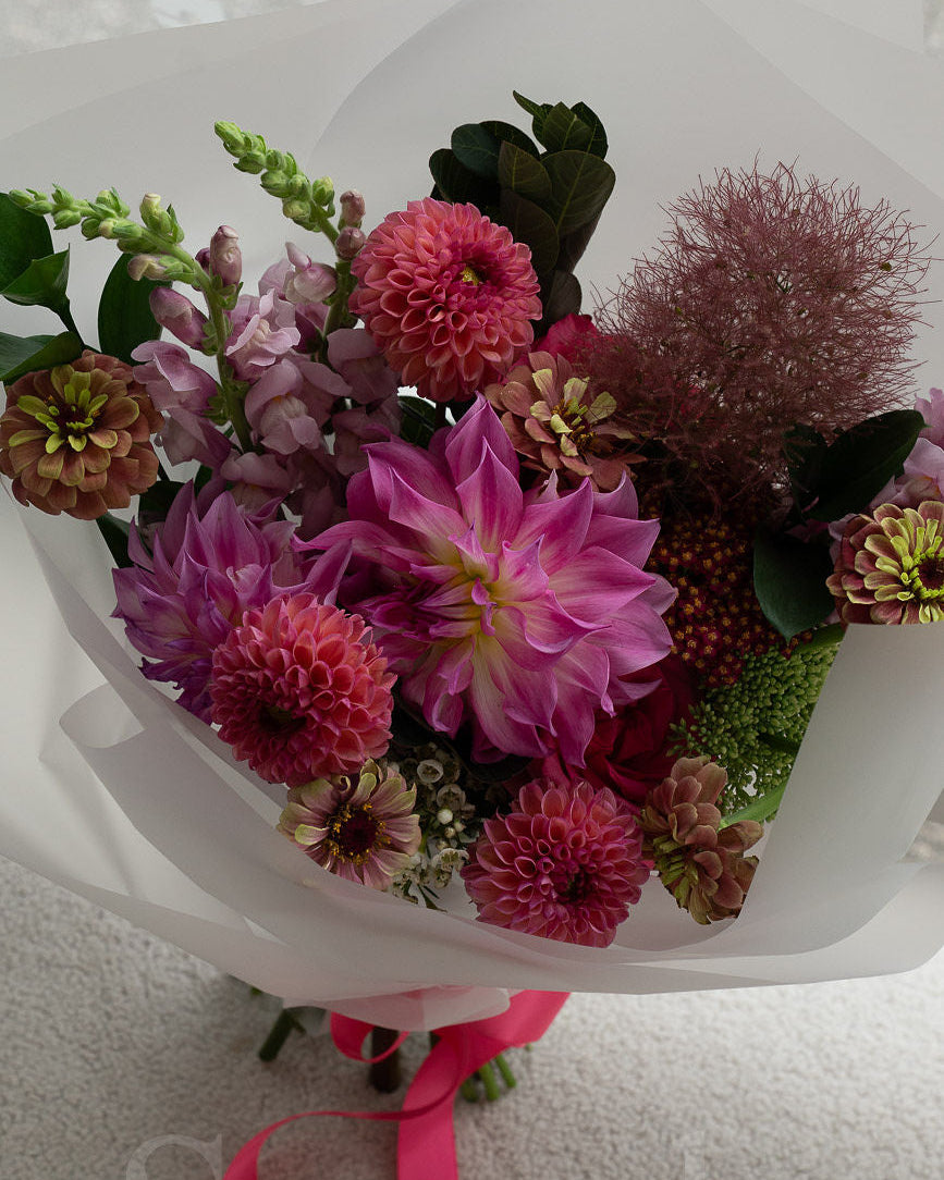 A vibrant flower bouquet featuring pink, peach and magenta dahlias, smoke bush, and pink and chartreuse zinnia flowers, wrapped in white paper with a bright pink ribbon, set against a neutral light gray background.