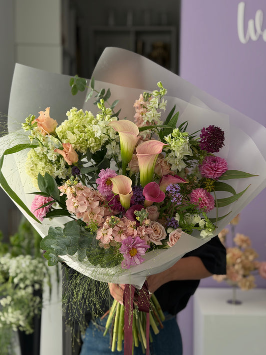 A large, soft and lush floral bouquet wrapped in white paper, held by a person in a shop. The bouquet features pink calla lilies, pink roses, purple dahlia, light green hydrangeas, and green foliage.