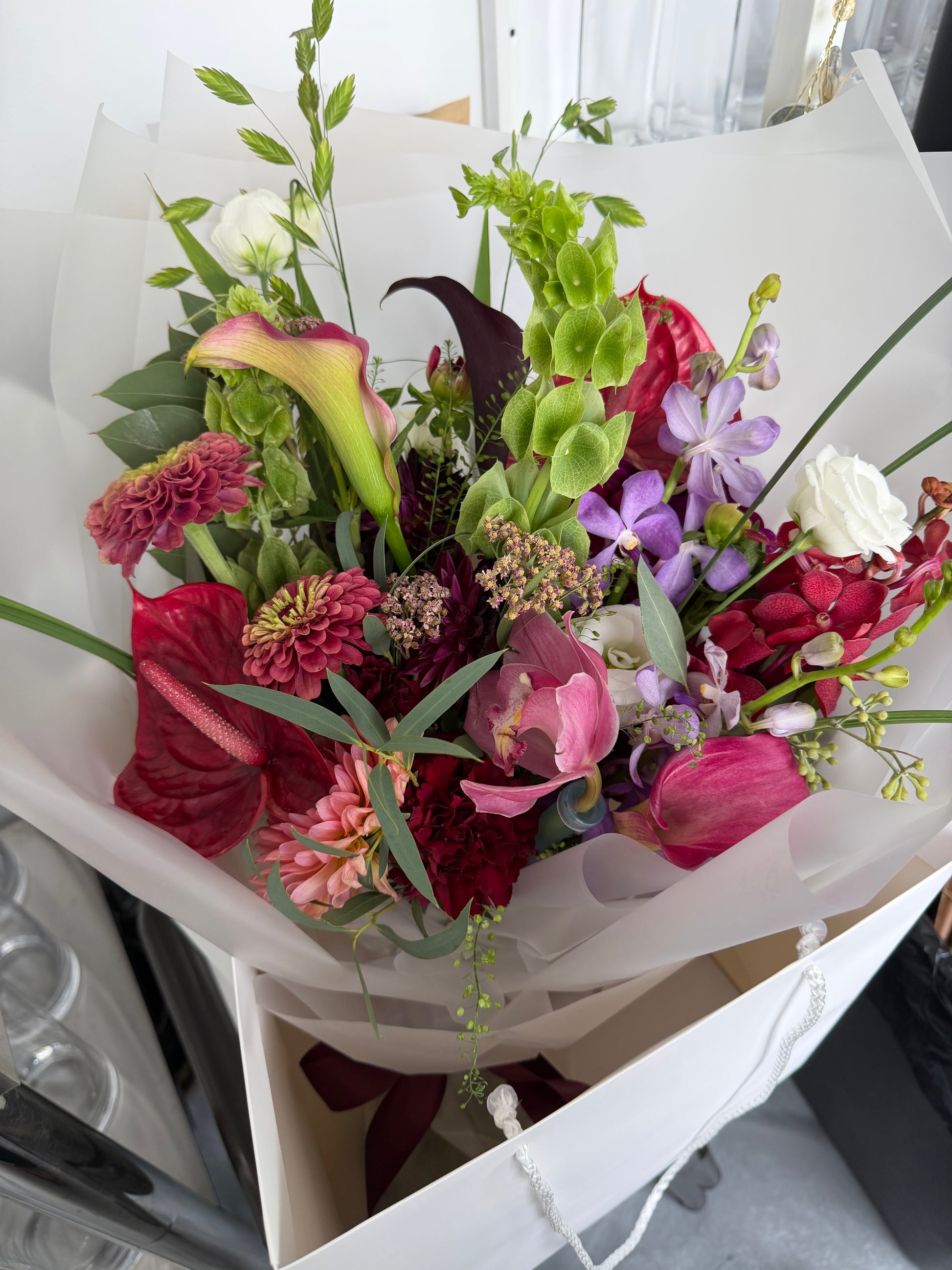 A vibrant mixed flower bouquet wrapped in white tissue paper and a white gift bag, featuring red anthuriums, pink and purple flowers, green foliage, and white roses.