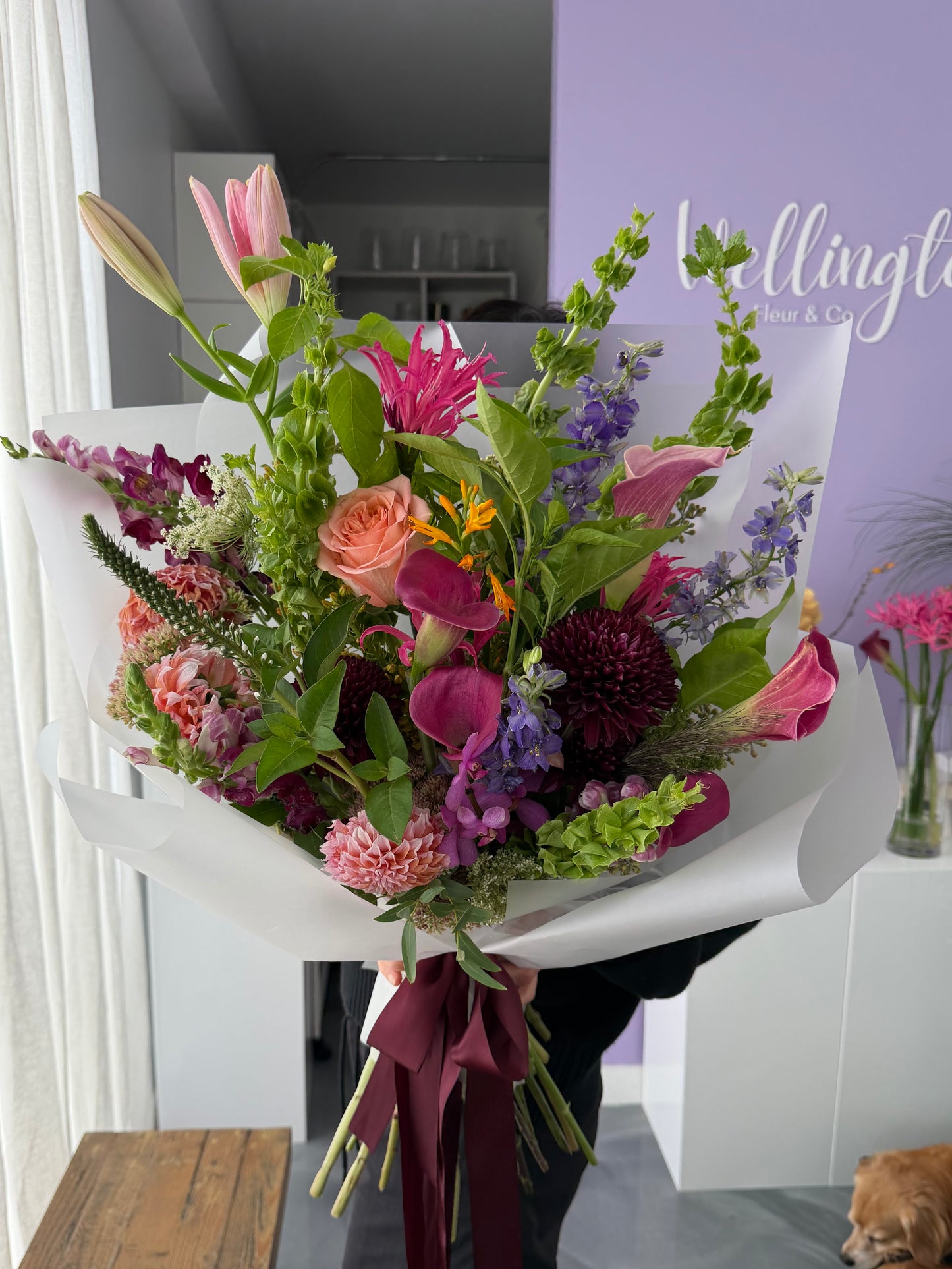A large, vibrant mixed flower bouquet held by a person in a shop setting. The bouquet features a variety of flowers including pink lilies, orange roses, purple calla lilies, burgundy dahlias, and green foliage, wrapped in white paper with a dark burgundy ribbon.