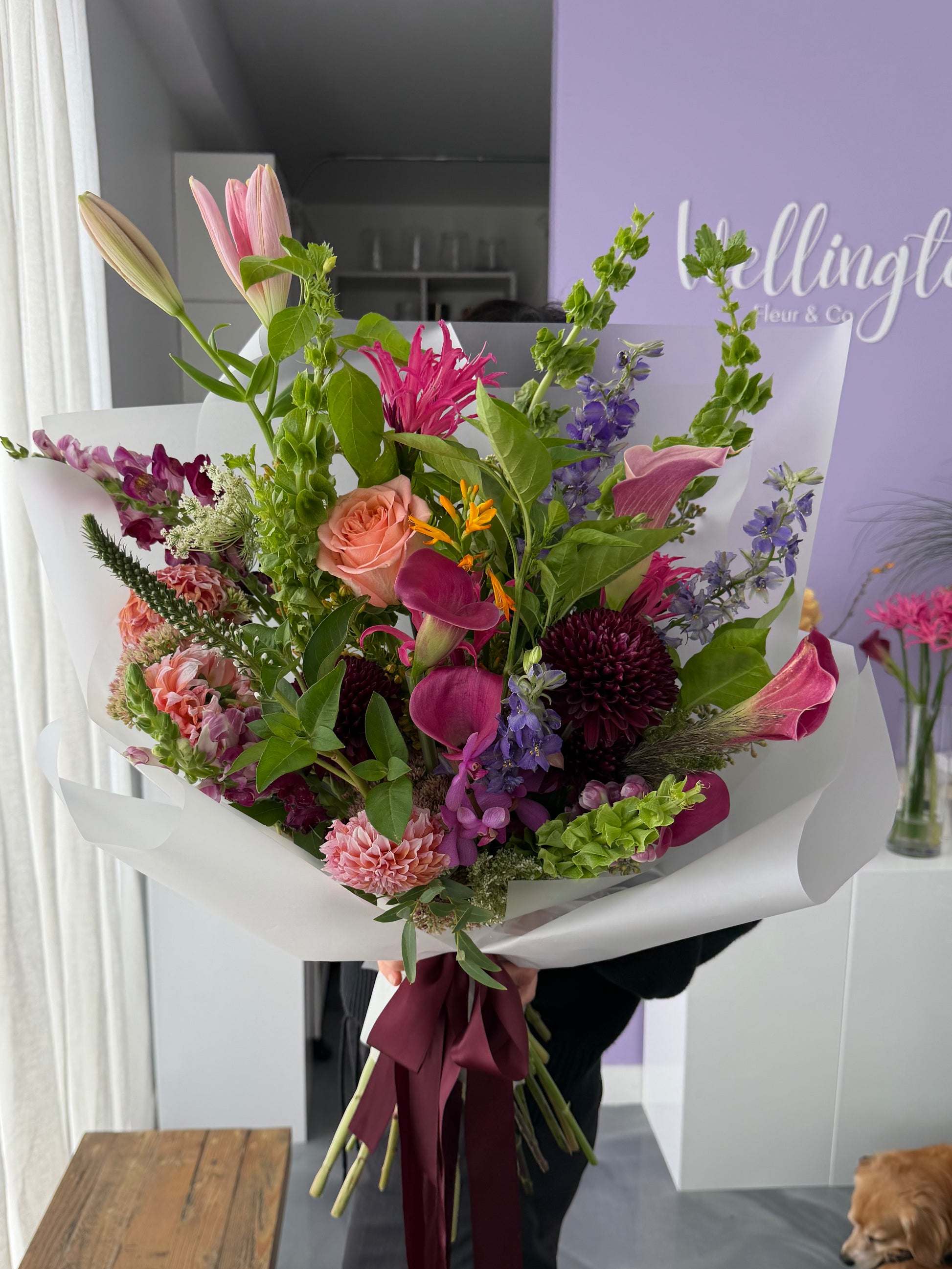 A large, vibrant mixed flower bouquet held by a person in a shop setting. The bouquet features a variety of flowers including pink lilies, orange roses, purple calla lilies, burgundy dahlias, and green foliage, wrapped in white paper with a dark burgundy ribbon.